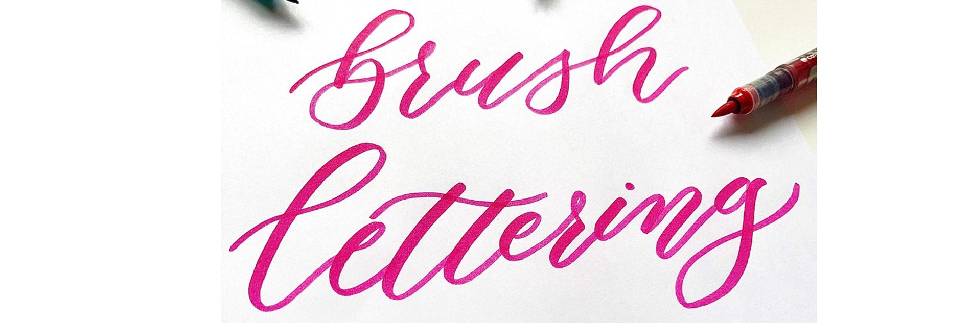 The phrase 'brush lettering' is depicted in bright pink ink on white paper. Three brush pens surround it, one green, one pink and one red.