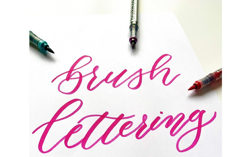 The phrase 'brush lettering' is depicted in bright pink ink on white paper. Three brush pens surround it, one green, one pink and one red.