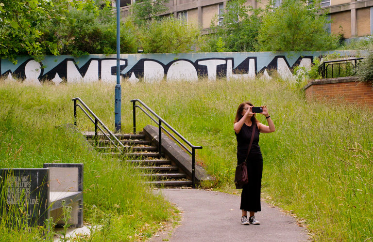 A photograph of an adult taking a photo on their phone. They are standing on a path with overgrown grass either side. Steps, a wall with painted street art and a large building can be be seen in the background.   