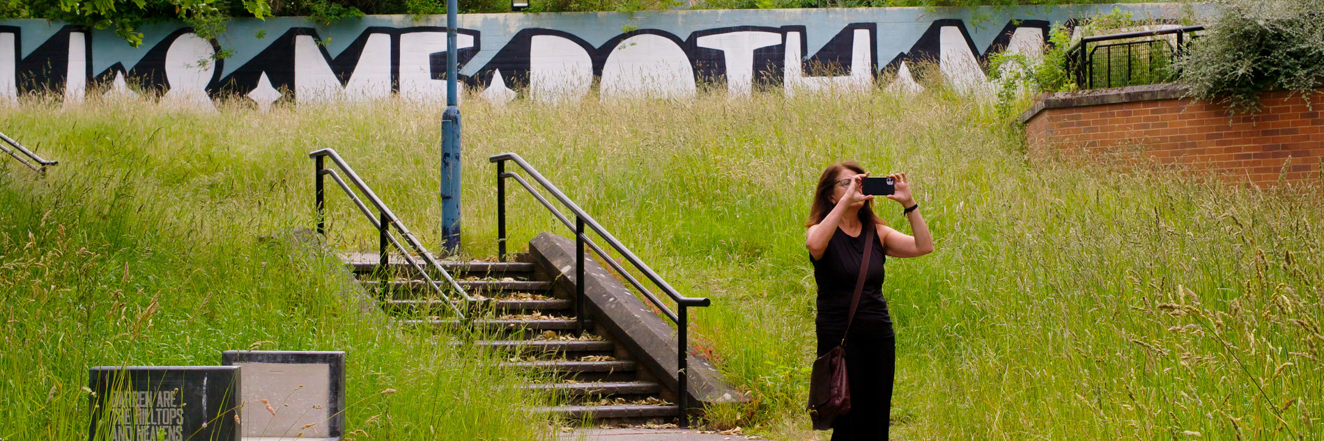 A photograph of an adult taking a photo on their phone. They are standing on a path with overgrown grass either side. Steps, a wall with painted street art and a large building can be be seen in the background.   