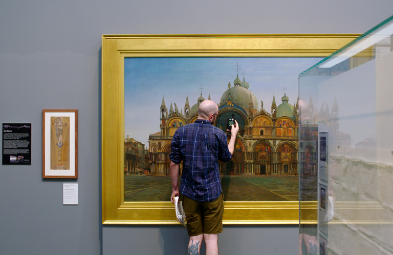 A photograph of an adult standing in front of a large painting in a gallery holding a small device and a clipboard. 