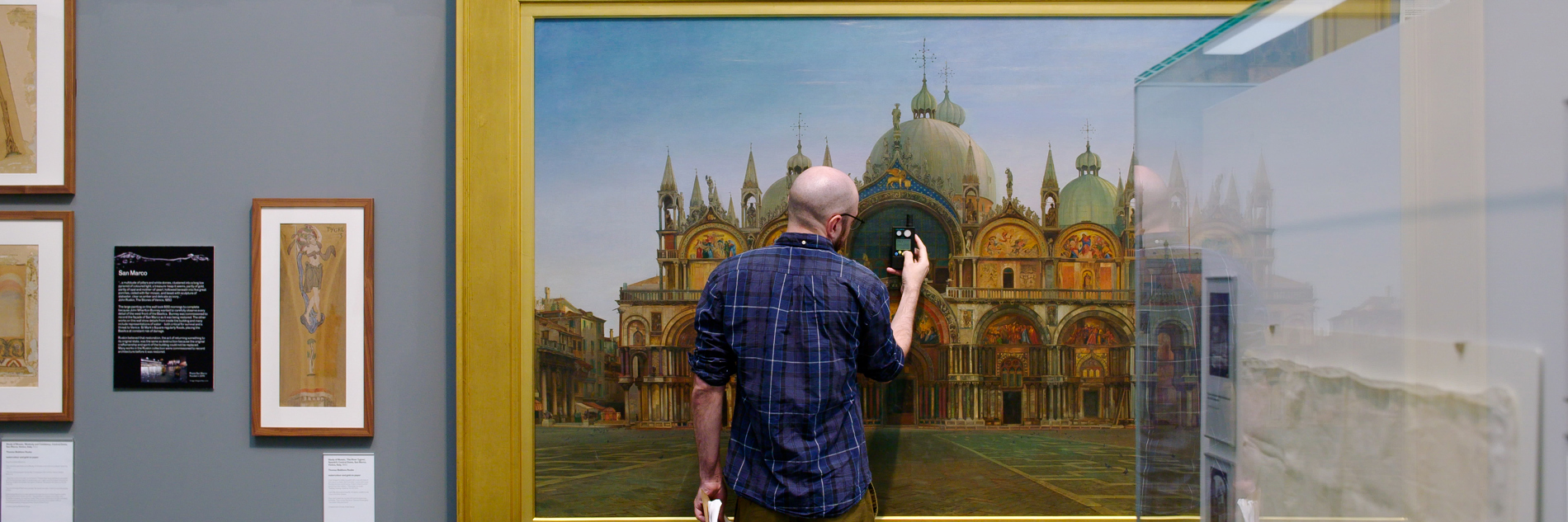 A photograph of an adult standing in front of a large painting in a gallery holding a small device and a clipboard. 