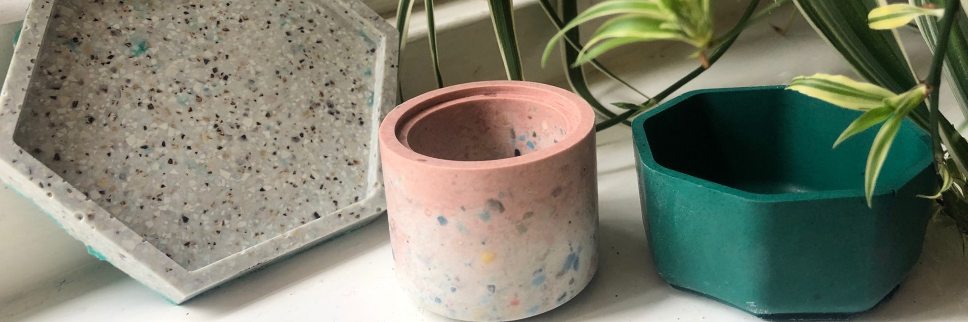 Three plant pots lined up on a windowsill with a houseplant partially visible in the top left corner. From left to right, a grey hexagonal plant pot saucer, a pink and white cylindrical plant pot and a green hexagonal plant pot. 