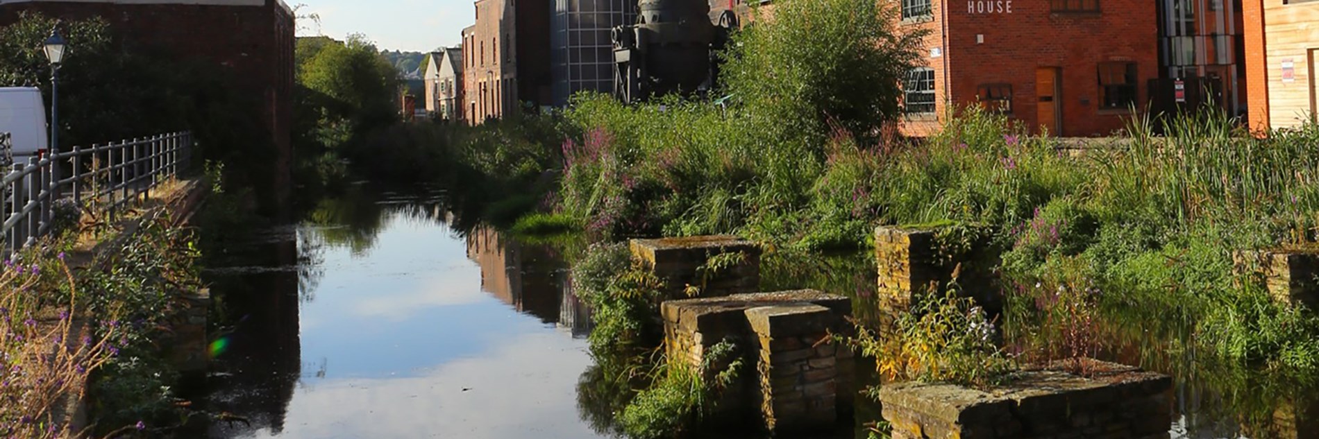 Photograph of a man-made water channel with greenery growing on the banks. To the side are red-bricked industrial buildings.