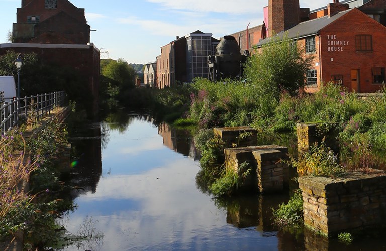 Photograph of a man-made water channel with greenery growing on the banks. To the side are red-bricked industrial buildings.