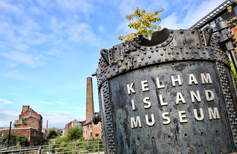 Huge metal industrial bucket with 'Kelham Island Museum' written on. Various buildings including one with a tall chimney are in the background.