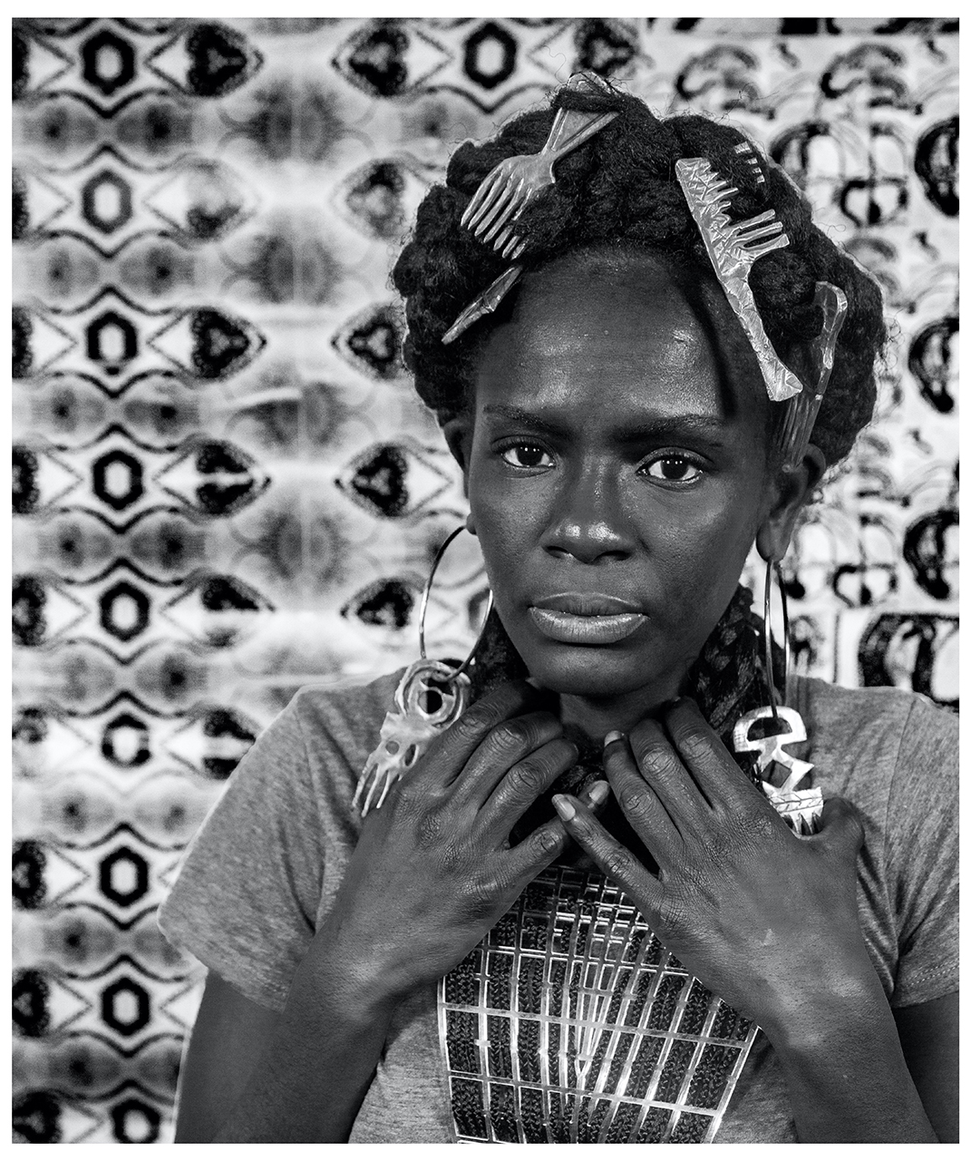Black and white photograph of a black woman with long, braided hair which comes around her neck to look like a necklace. She is holding both hands to her neck and is also wearing large, comb-shaped, hair accessories and hoop earrings. She is looking directly at the camera with a serious expression and stands in front of a patterned background.