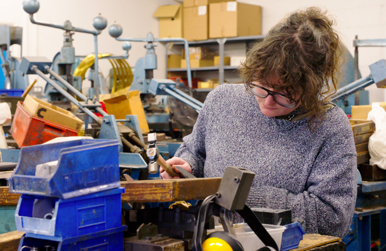A photograph of an adult in a workshop, using a small hammer on a piece of wood. Various plastic crates are stacked in front of them. 