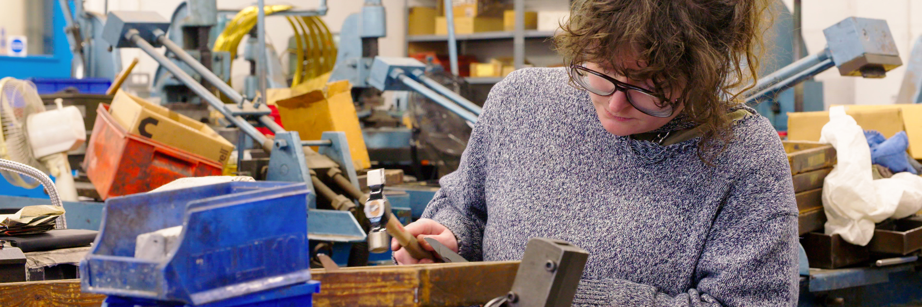 A photograph of an adult in a workshop, using a small hammer on a piece of wood. Various plastic crates are stacked in front of them. 