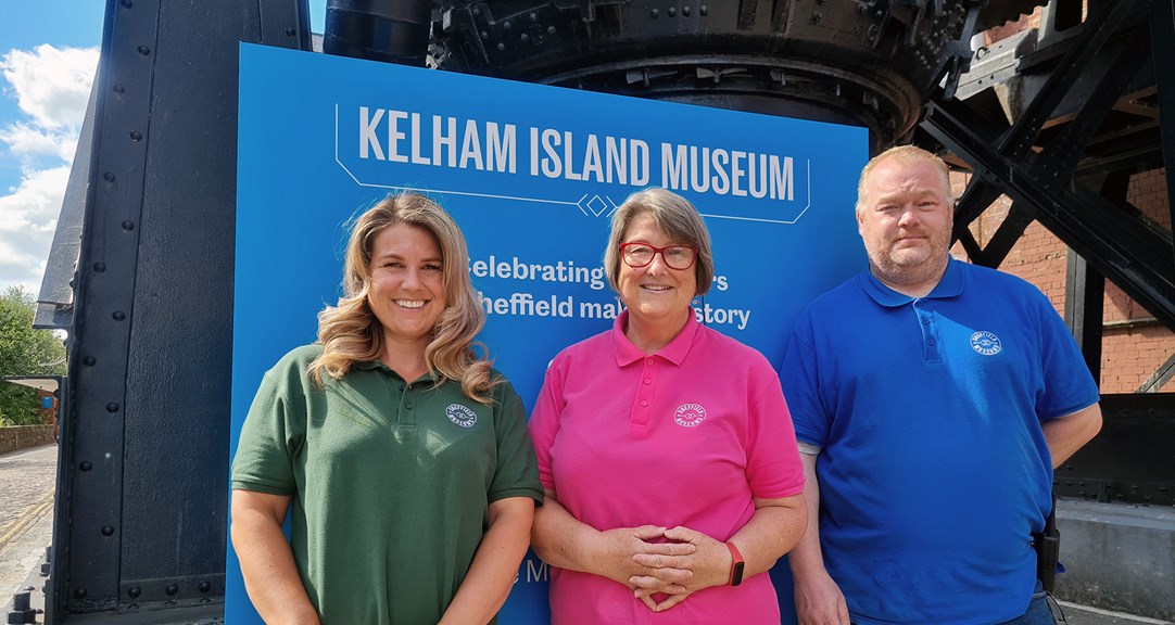 Three adults in pink, blue and green polo shirts with the Sheffield Museums logo in front of a sign promoting Kelham Island Museum