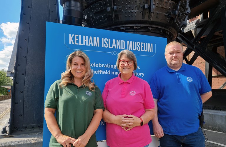 Three adults in pink, blue and green polo shirts with the Sheffield Museums logo in front of a sign promoting Kelham Island Museum