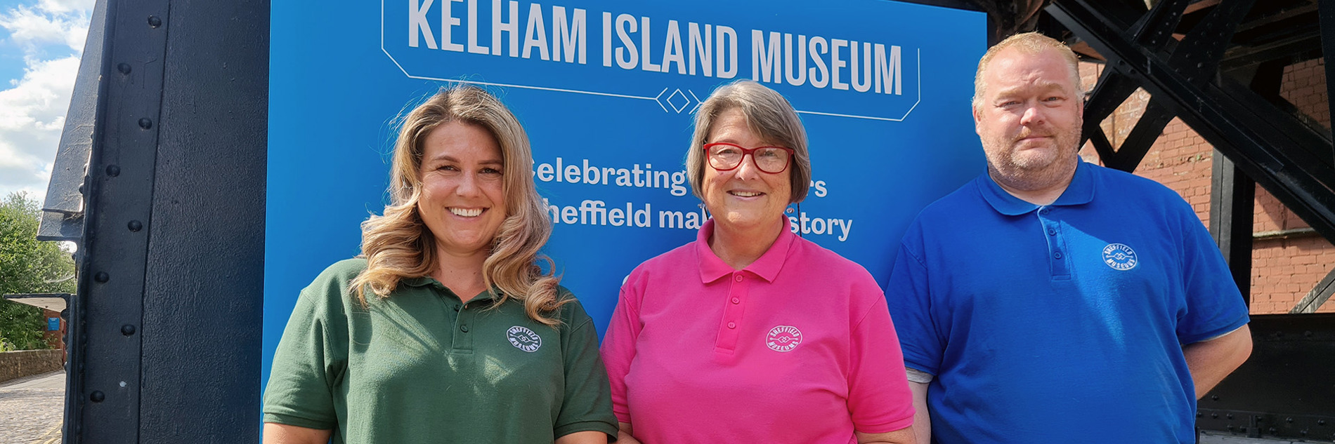 Three adults in pink, blue and green polo shirts with the Sheffield Museums logo in front of a sign promoting Kelham Island Museum
