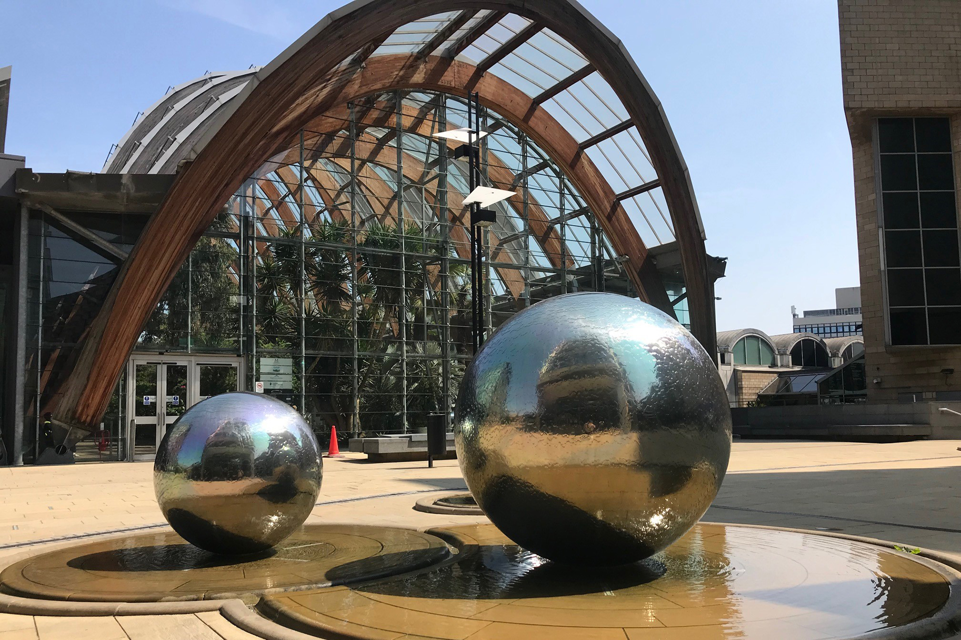 Photograph of 2 large silver spheres sitting in shallow pools of water on a sunny day. In the background there is a large wooden and glass arched structure filled with plants.