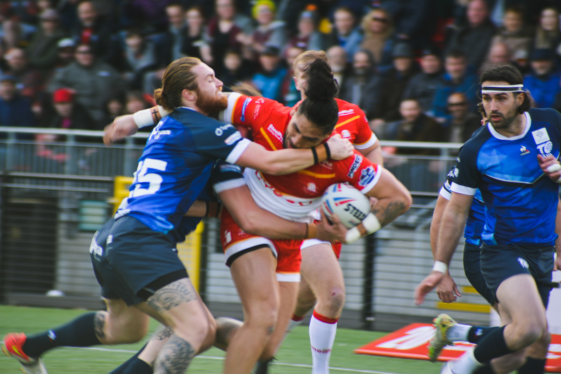 A group of adults playing rugby in red and blue rugby kits. A crowd of spectators can be seen in the background. 