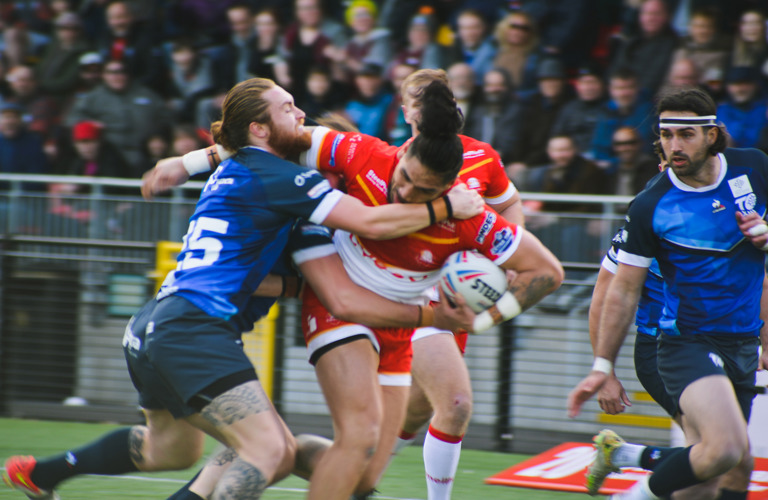 A group of adults playing rugby in red and blue rugby kits. A crowd of spectators can be seen in the background. 