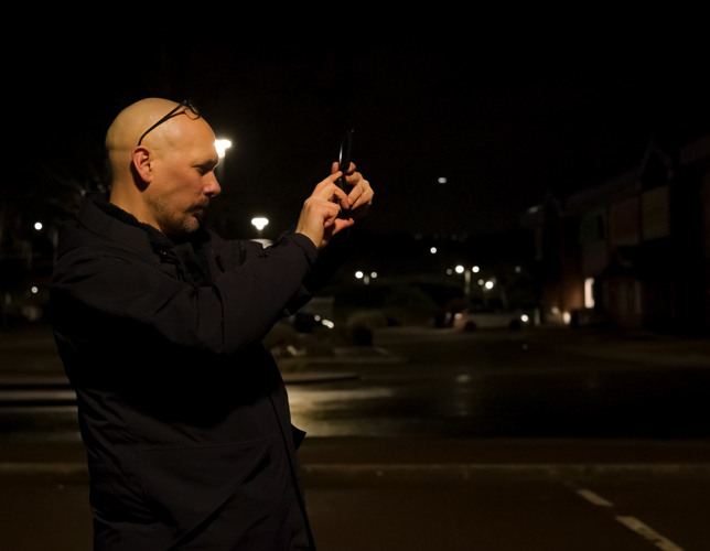 An adult taking a photograph at night. In the background there is a residential street, street lights and cars. 
