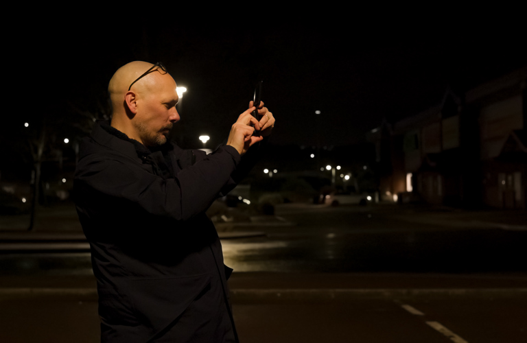 An adult taking a photograph at night. In the background there is a residential street, street lights and cars. 