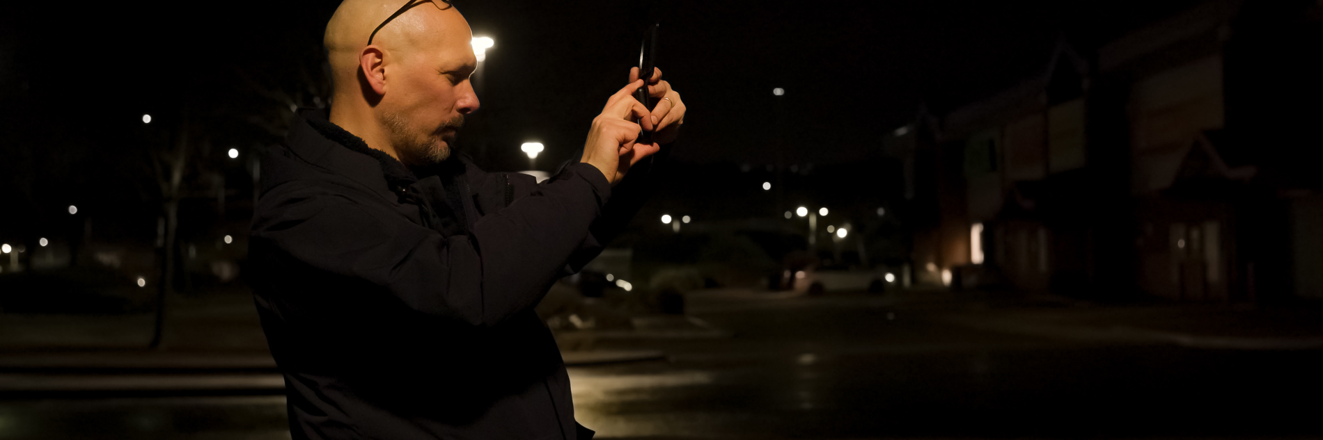 An adult taking a photograph at night. In the background there is a residential street, street lights and cars. 