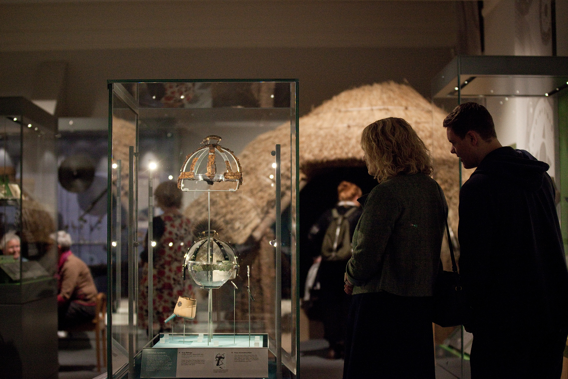 Two adults stood looking at a display case with the remains of a helmet with a boar on top - there is a modern replica below the original. The background is a exhibition area with visitors and other displays including the Iron Age Roundhouse.