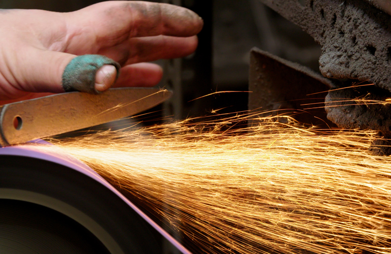 A close-up photograph of a hand of a person holding a knife on a grinding wheel and creating sparks. The person's fingers on their hand are dirty and they have a plaster on their thumb. 