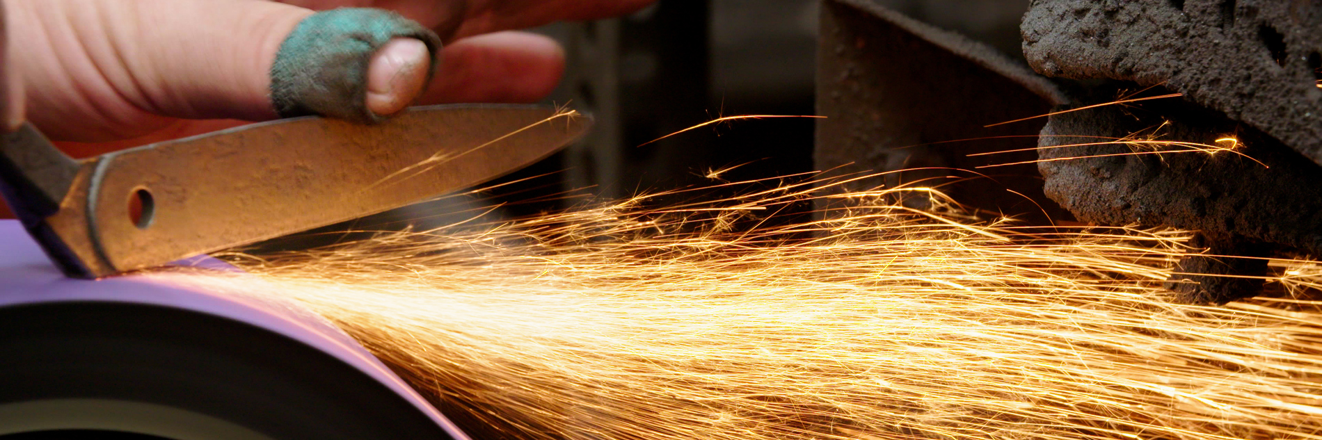 A close-up photograph of a hand of a person holding a knife on a grinding wheel and creating sparks. The person's fingers on their hand are dirty and they have a plaster on their thumb. 