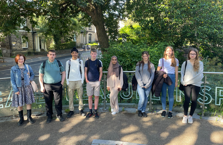 6 young people standing on the path in front of Weston Park Museum. Behind them is a waist high glass barrier with a metal hand rail. Behind that are bushes and trees, with the road and buildings visible in the background to the left of the image. 