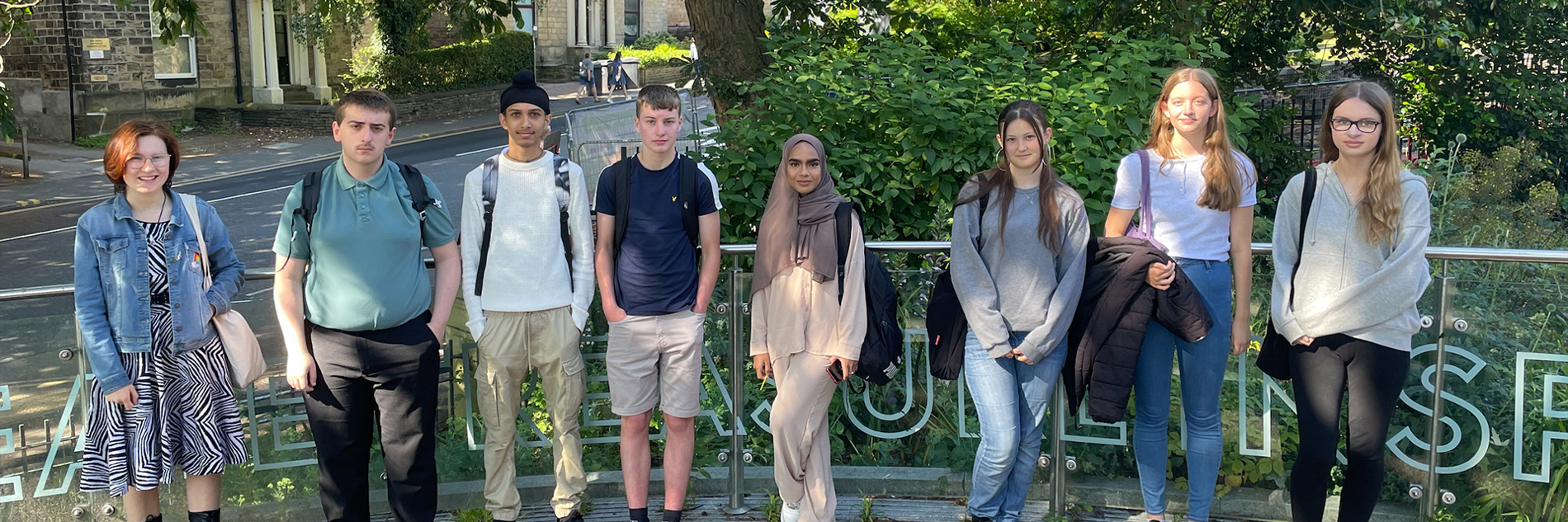 6 young people standing on the path in front of Weston Park Museum. Behind them is a waist high glass barrier with a metal hand rail. Behind that are bushes and trees, with the road and buildings visible in the background to the left of the image. 