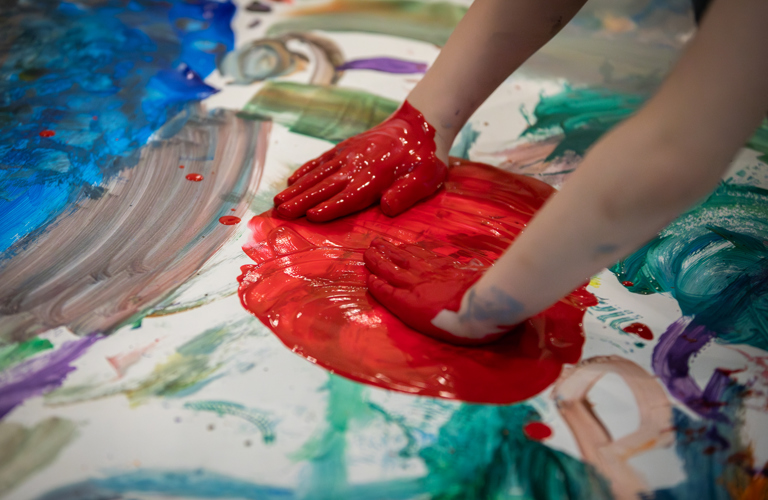 A photograph of a child's hands smearing a big red circle of paint on paper, on top of other colours.