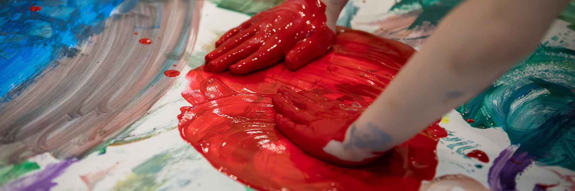 A photograph of a child's hands smearing a big red circle of paint on paper, on top of other colours.