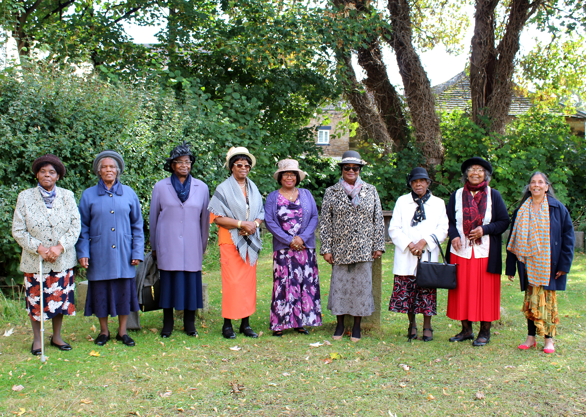 A photograph of the Black Ladies Group. There are eight people standing in a line, some are wearing hats. They are outside with trees behind them.