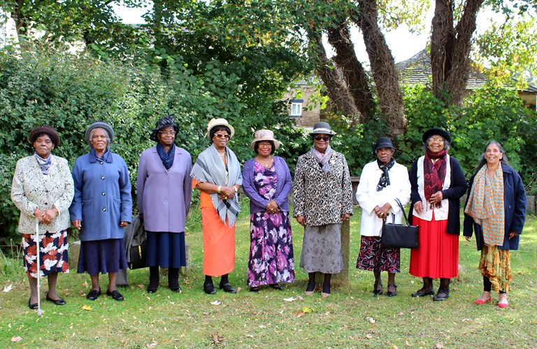 A photograph of the Black Ladies Group. There are eight people standing in a line, some are wearing hats. They are outside with trees behind them.
