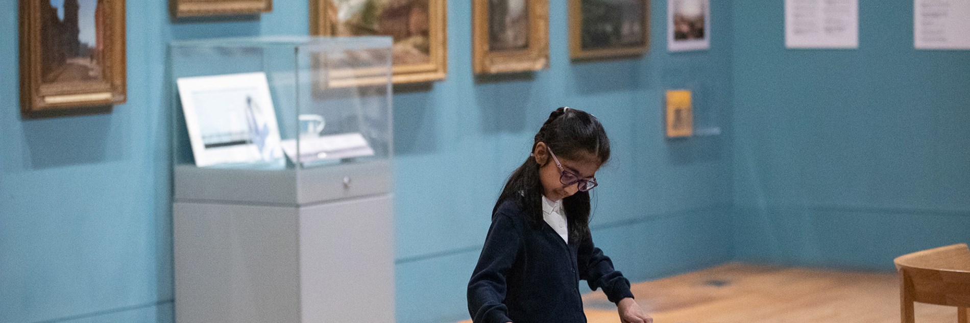 A primary school student leans over a table in a gallery with framed paintings on the walls. 