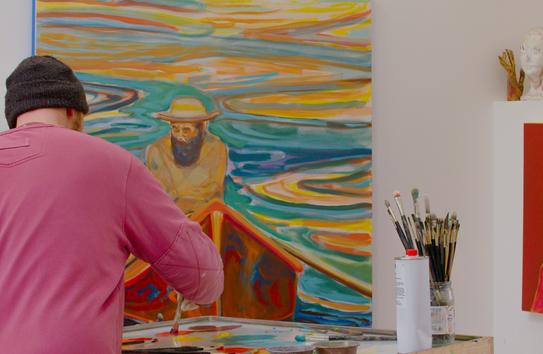 A photograph of an adult stood painting with their back to the camera. In front of them is a paint palette, art supplies and paint brushes. In the background there is a large painting, a smaller painting and a collection of sculptures.  