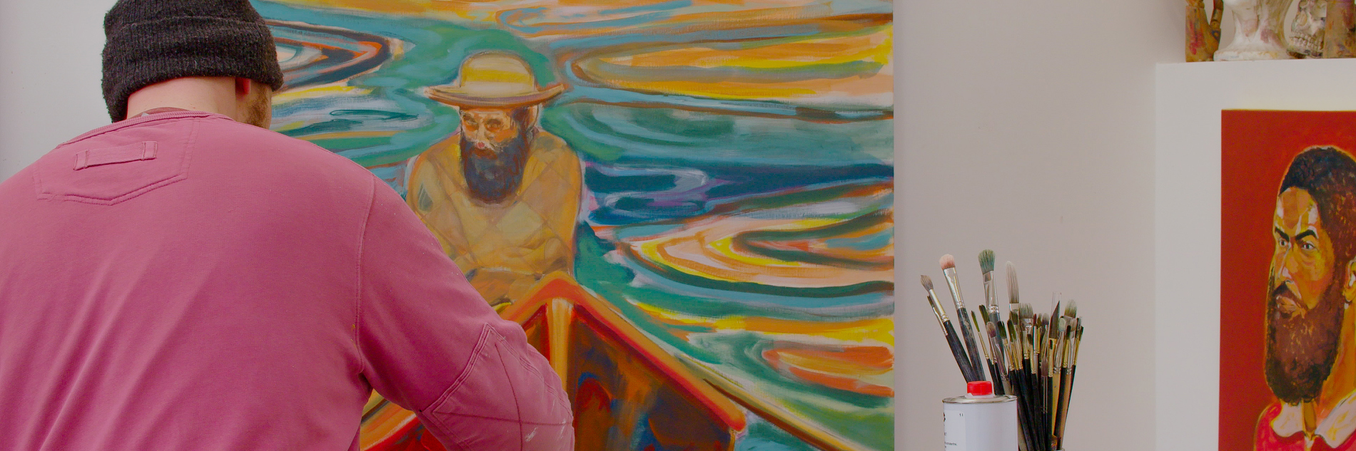 A photograph of an adult stood painting with their back to the camera. In front of them is a paint palette, art supplies and paint brushes. In the background there is a large painting, a smaller painting and a collection of sculptures.  