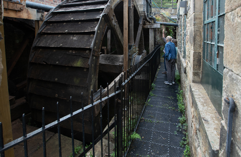 Visitors on a metal walkway next to a large, historic wooden waterwheel