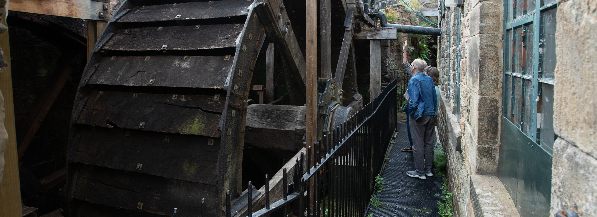 Visitors on a metal walkway next to a large, historic wooden waterwheel