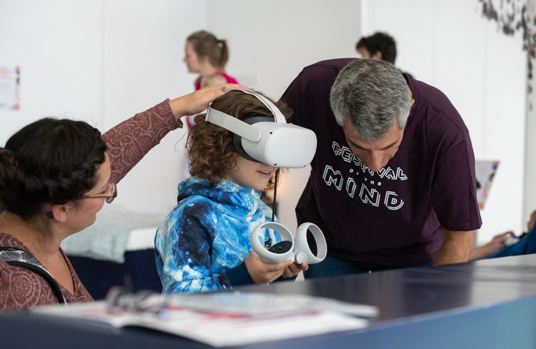 A photograph of a child and two adults in an exhibition. The child is wearing a virtual reality headset and holding two controllers. 