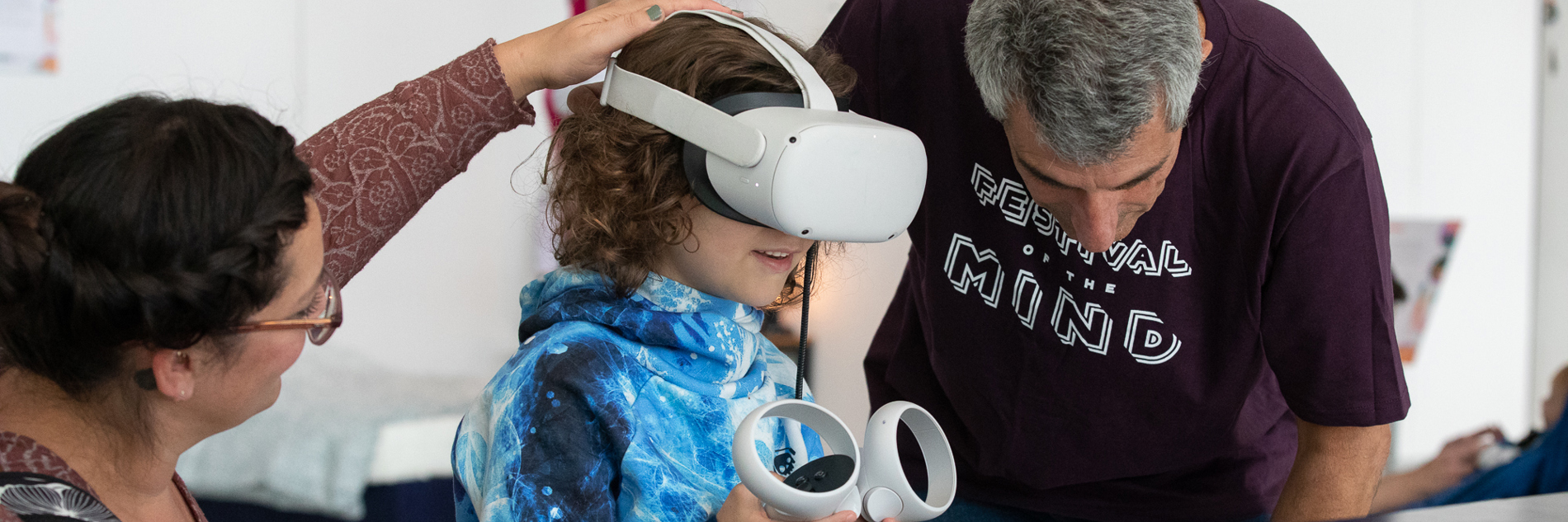 A photograph of a child and two adults in an exhibition. The child is wearing a virtual reality headset and holding two controllers. 