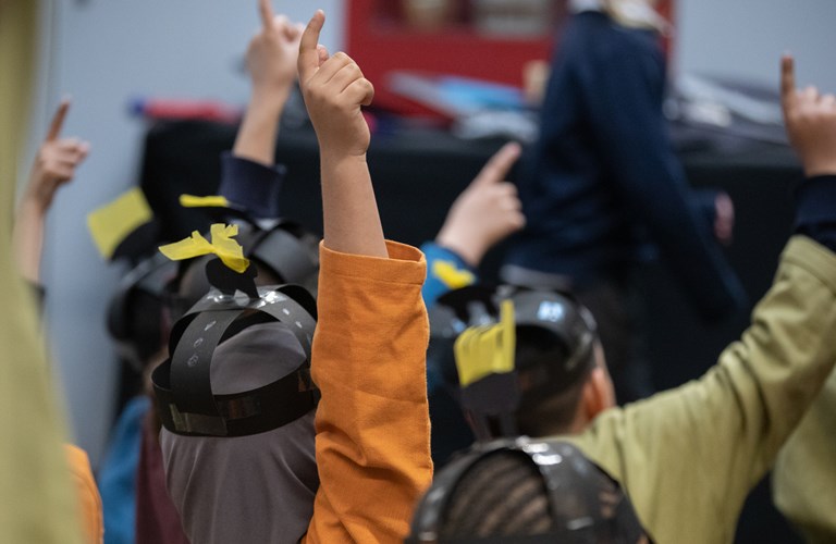 A view from behind of several primary school aged children with raised hands. They are dressed as Anglo Saxons and wearing brightly coloured tunics and paper helmets. 