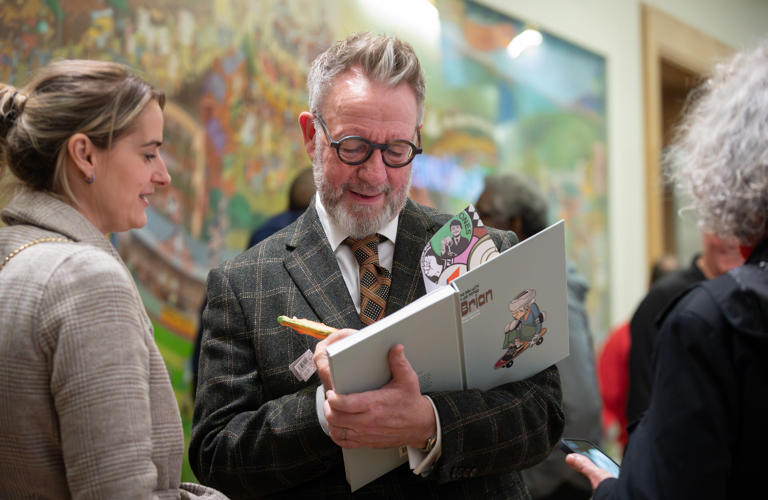 A photograph of an adult signing a book. An adult is standing next to them and other adults can be seen in the background. 