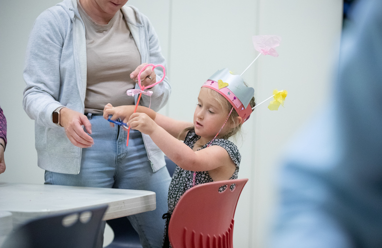 A photograph of a young girl in a colourful homemade pink party hat. She is shown using scissors to cut a pipe cleaner, probably to use as a magic wand.
