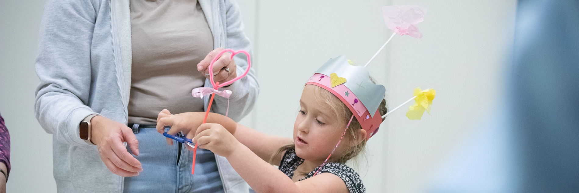 A photograph of a young girl in a colourful homemade pink party hat. She is shown using scissors to cut a pipe cleaner, probably to use as a magic wand.